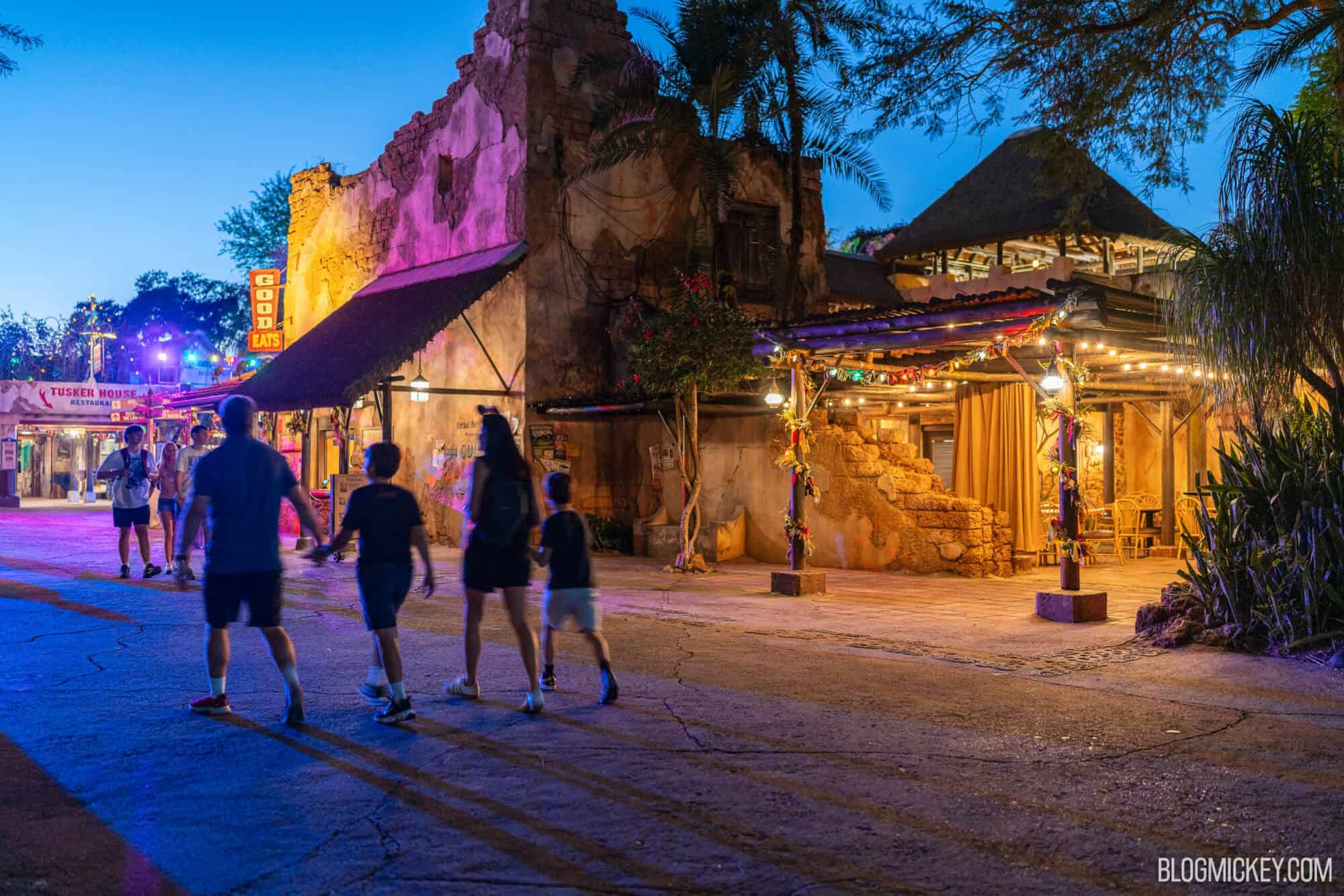Vibrant outdoor scene at dusk with visitors walking past themed rustic buildings and decorative lights at Disney World.