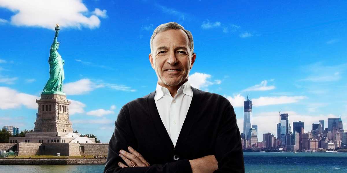 A man with gray hair and a light smile stands with his arms crossed in front of the Statue of Liberty and the New York city skyline on a sunny day with a clear blue sky. He is wearing a white shirt and a black sweater.