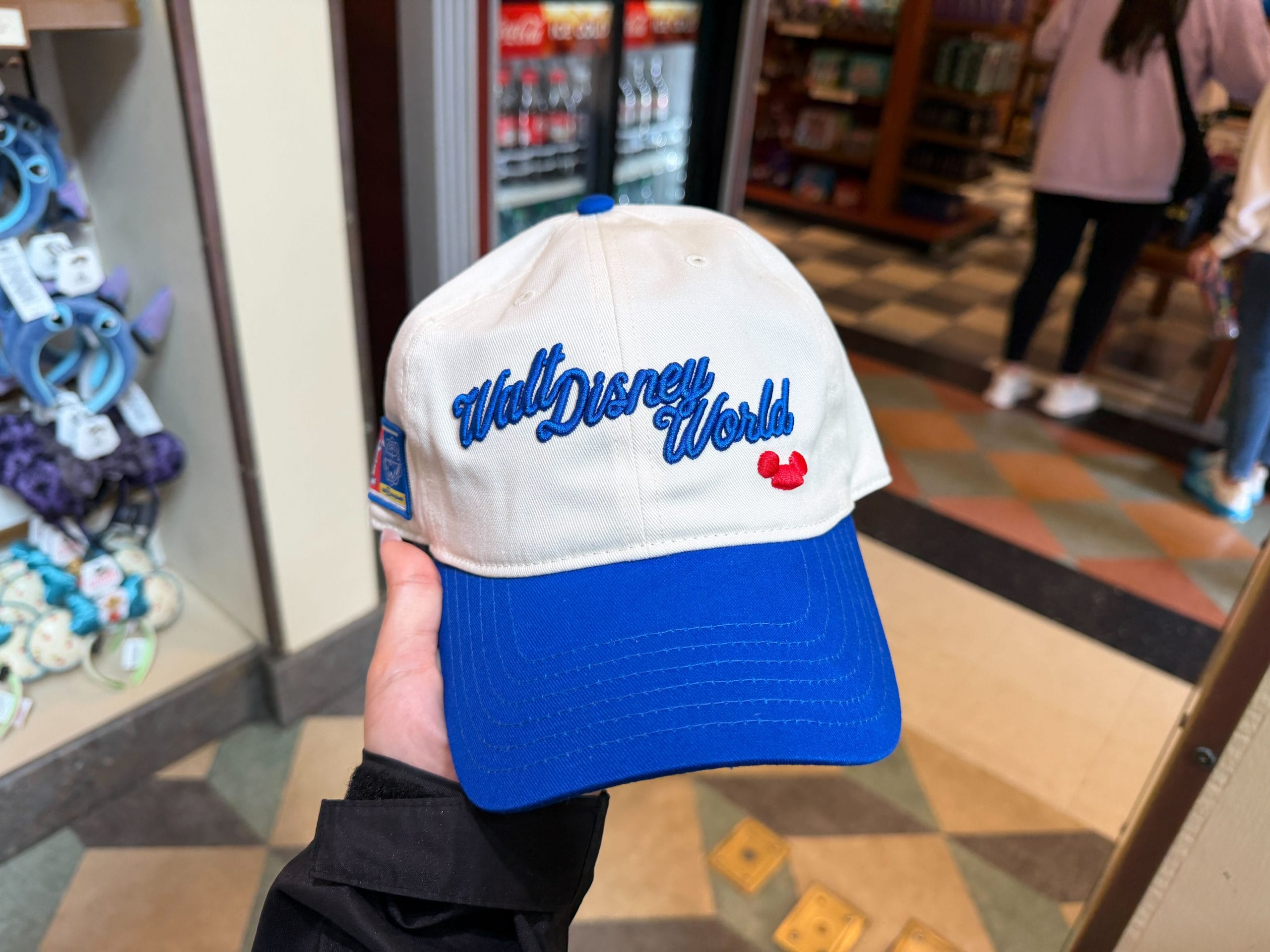 This white baseball cap has Walt Disney World in royal blue script on the front. The bill and button on top of the hat are in a matching blue. There is also a red embroidery of a Mickey ear hat on the bottom front of the hat.