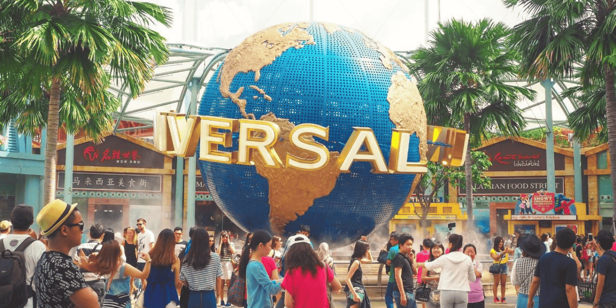 Crowd of people at an amusement park entrance with a large globe sculpture labeled "Universal" overhead, surrounded by colorful buildings and palm trees within Universal Studios Hollywood with the Jurassic World ride close by at Universal Studios Hollywood.
